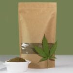 Kraft paper packaging with a cannabis product inside and a cannabis leaf and ramekin of product leaning against the package.