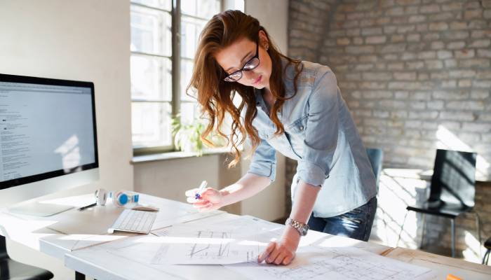 A young female architect looks over building plans scattered across her desk. She has a blue marker in her hand.