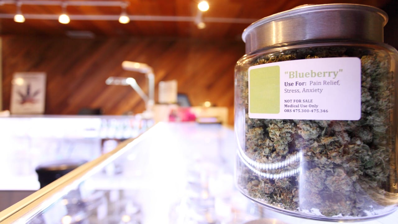 A glass jar with a silver lid contains marijuana buds. It has a label that tells the customer the type of weed it is.