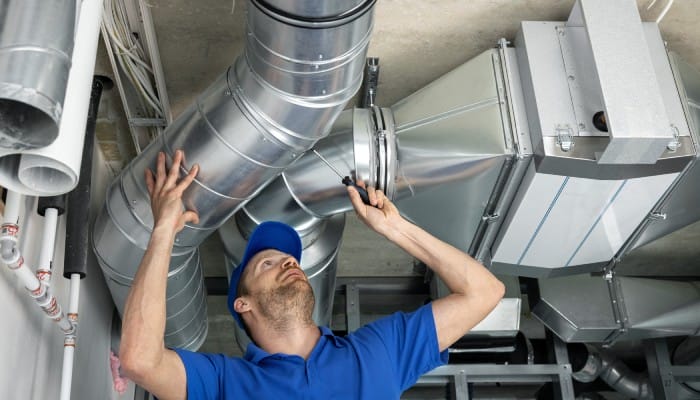 An HVAC technician in a blue shirt and hat using a screwdriver to perform maintenance on a facility’s HVAC system.