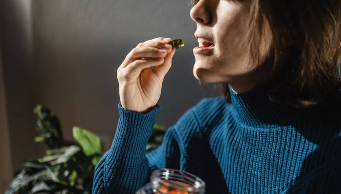 A woman in a blue sweater holding a jar of colorful CBD gummies and preparing to place one in her mouth.