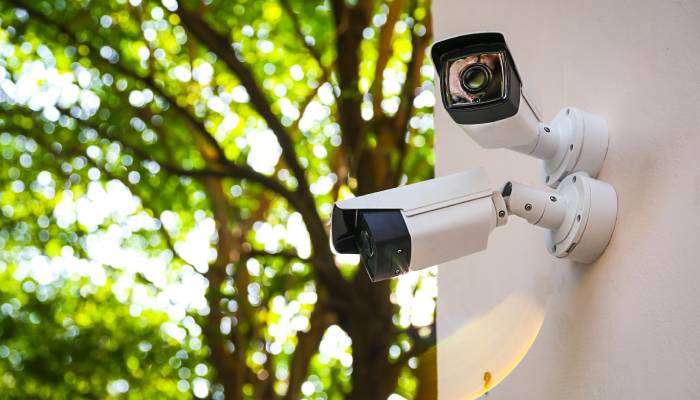 Two security cameras hang on a white exterior wall. One camera points to the left, while the other points to the right.