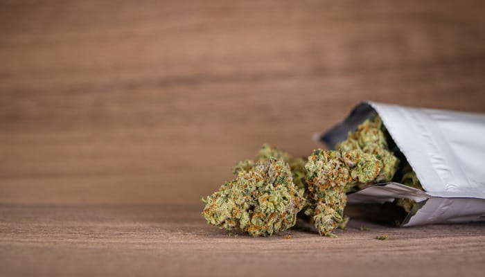 A silver ziplock bag sits open on a wooden surface, and marijuana buds are spilling out the opening.