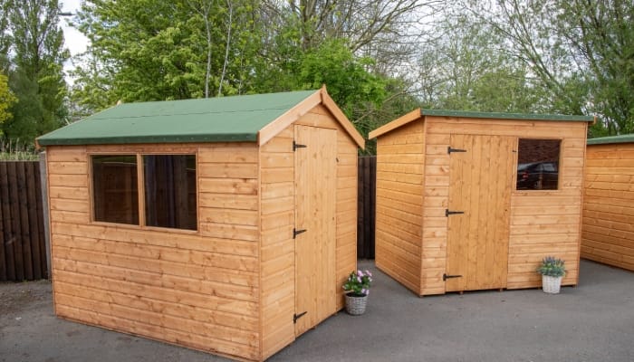 Two wooden sheds on paved ground with green roofs. They each feature a door and window and there's plants outside the door.