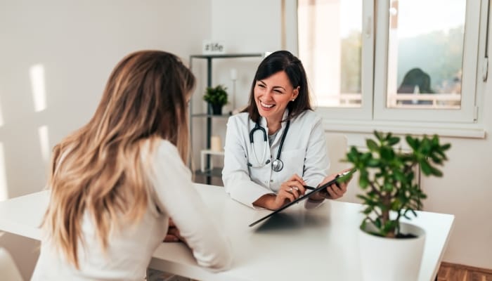 A doctor sits at a desk and points to a clipboard while smiling at a patient. The patient's back is toward the camera.