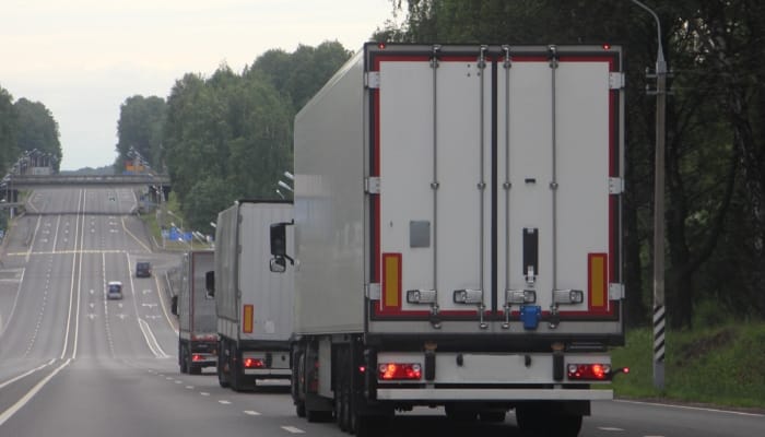 Three tractor-trailers drive down the road, surrounded by mature trees in a line. Each has a white trailer.