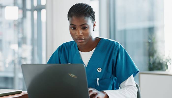 A woman wears a white long-sleeve shirt under her teal scrubs as she sits at a desk looking at an open silver laptop.
