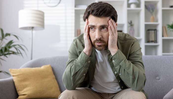 A worried middle-aged man sitting on the edge of a couch with his hands pressed against the sides of his face.