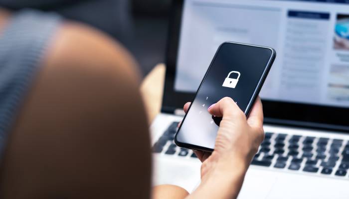 A woman sits in front of an open laptop while holding a cell phone showing a lock symbol on the screen.