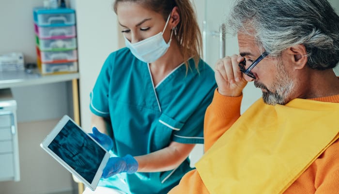 A female dental professional wearing a blue mask shows an x-ray of teeth to a male patient wearing a yellow sweater.