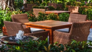 Home 20 A square wooden table with four large, brown chairs sits beside a bubbling fountain in a pot and plants.