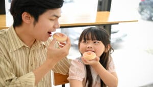 A father and his young daughter sit in wooden chairs enjoying cake doughnuts with sugar on top. They are taking a bite.