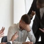 A man sits at a table with his head in his hand while four people point their fingers at him or yell at him.