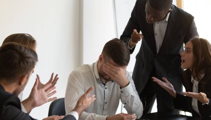 A man sits at a table with his head in his hand while four people point their fingers at him or yell at him.