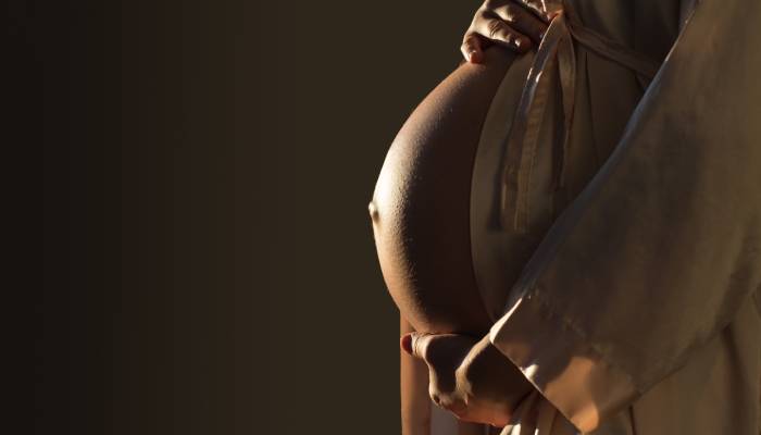 A dimly lit and warm side portrait of a pregnant person’s bare belly. They are wearing a beige robe.