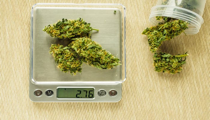 Three large cannabis buds resting on a silver digital scale that reads 2.76 grams. To the right is a plastic jar of more buds.