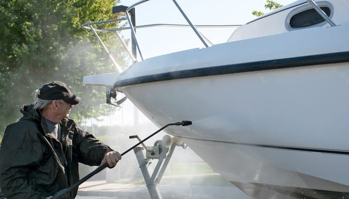 A man wearing a black jacket and black baseball cap uses a pressure washer on a large, white boat.