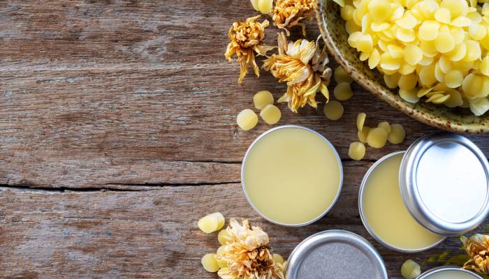 Several small tins filled with light yellow balm sitting on a wooden board alongside golden flowers and wax balls.