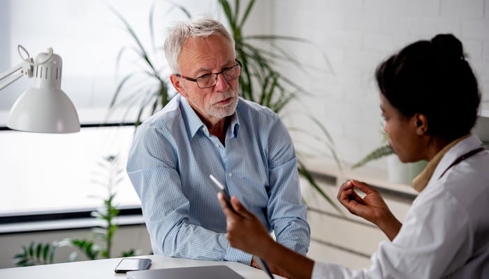 An older man sitting in a bright doctor’s office and reviewing the information a medical professional is sharing with him.