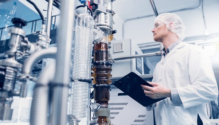 A lab tech looking at a machine extracting CBD oil. He's wearing protective gear and holding a clipboard.