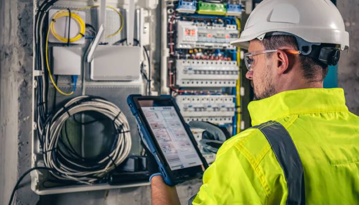 A technician in a hard hat and yellow uniform works with an electrical fuse box. He holds a tablet in one hand.
