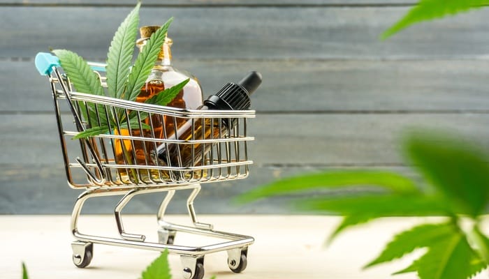 A miniature metal shopping cart filled with a cannabis leaf and two glass bottles of tinctures against a wooden background.