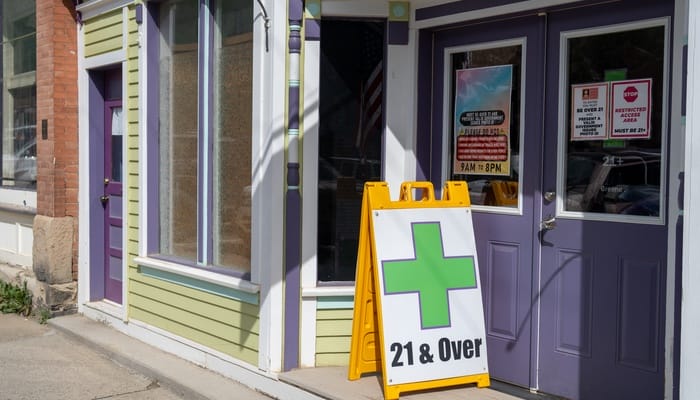 The exterior of a cannabis dispensary with a curb sign that has a green plus sign on it signaling the business is open.