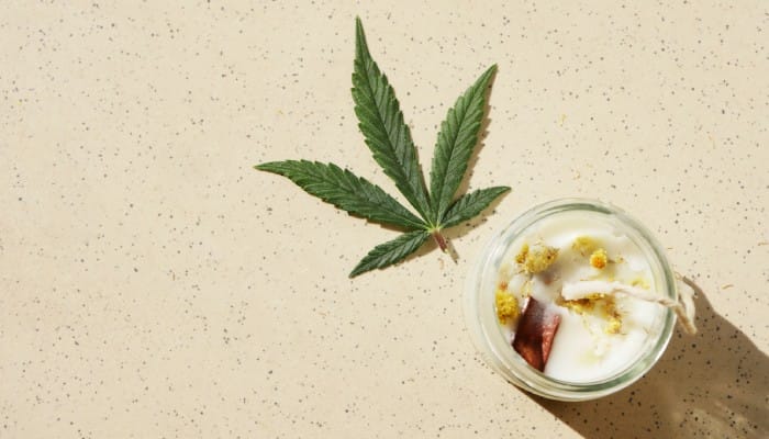 A top view of a marijuana leaf and a CBD candle in a glass jar resting on a speckled beige surface.