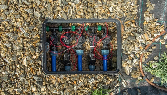 A ground-level irrigation valve box holding wires and pipe connections while sitting among wood chips and landscaping fabric.