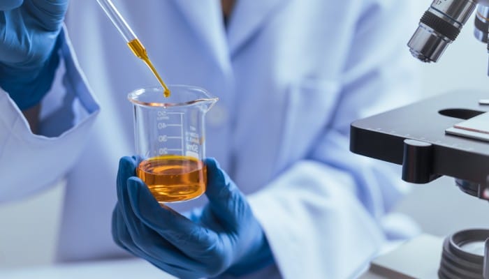 A person wearing a white lab coat and blue gloves using a dropper to drip an orange liquid into a beaker.
