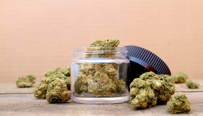 A small glass jar of marijuana buds with the lid off, sitting on concrete against a light orange wall.