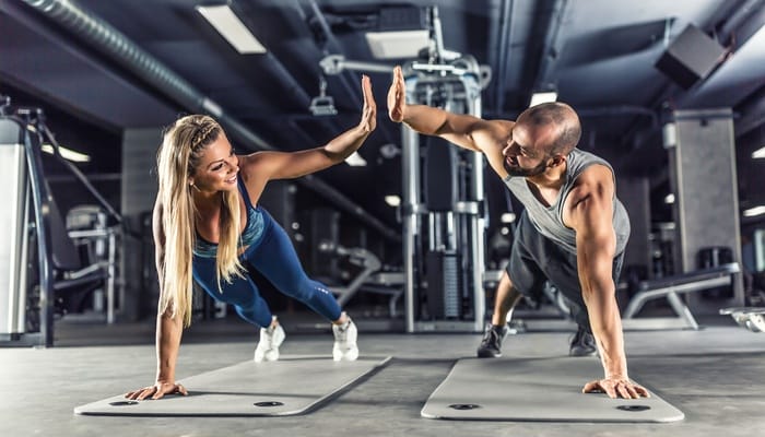 A man and woman are doing planks on gym mats and giving each other a high five. Machines are in the background.