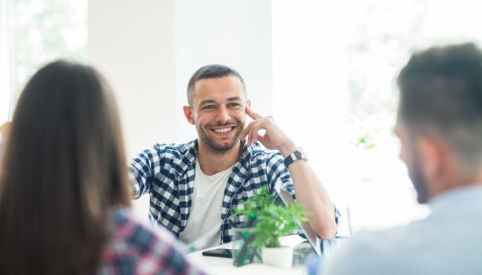 A man and a woman are sitting across a desk from a young man wearing a plaid shirt who is smiling widely.