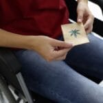 A person sitting in a standard wheelchair and holding a small brown bag in both hands. The bag has a cannabis leaf on it.