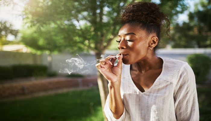 A woman with her hair up, standing outside near a tree and inhaling smoke from a rolled marijuana cigarette.