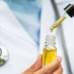A professional in a white lab coat lifting a full pipette from a bottle of translucent yellow CBD oil.
