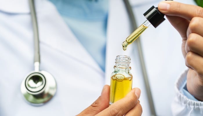 A professional in a white lab coat lifting a full pipette from a bottle of translucent yellow CBD oil.
