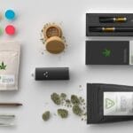 A selection of cannabis products, including a vape cartridge, grinder, and electronic cigarette, laid out on a white surface.