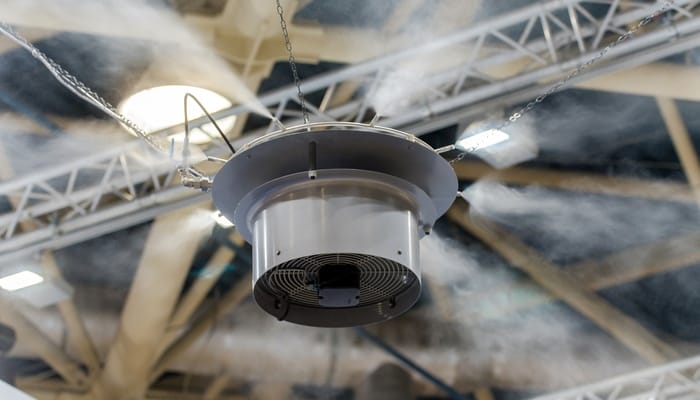 A circular industrial swamp cooler strung to a building's ceiling beams with three chains. The cooler emits mist.