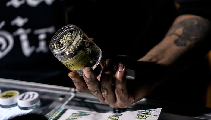 A person with arm tattoos is holding a small jar of cannabis over a glass countertop covered in paper.