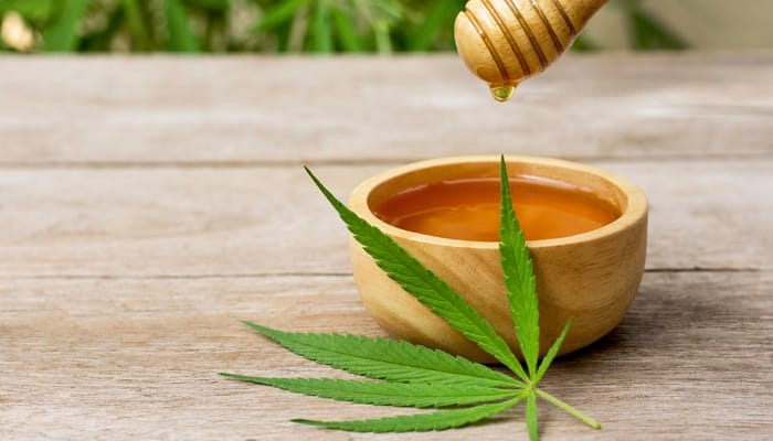 A bowl of honey with a honey-covered spool hanging over it. There's also a cannabis leaf sitting against the bowl.