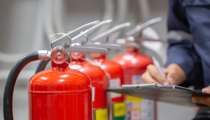 A close-up shows a person holding a clipboard and a pen next to a line of four red fire extinguishers.
