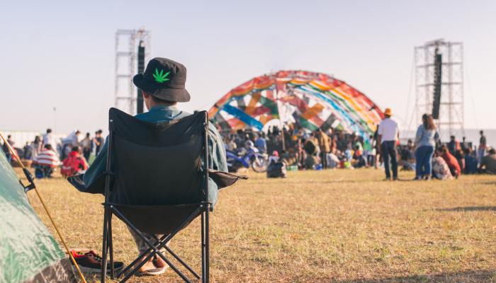 A person sits in a black camping chair with a black cannabis bucket hat on. They face the outdoor event stage.
