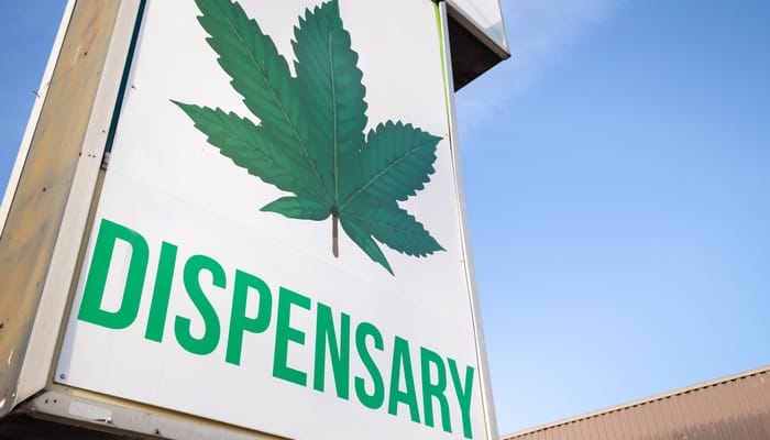 A sign in front of a building featuring a marijuana leaf and the word "DISPENSARY" written below in green letters.