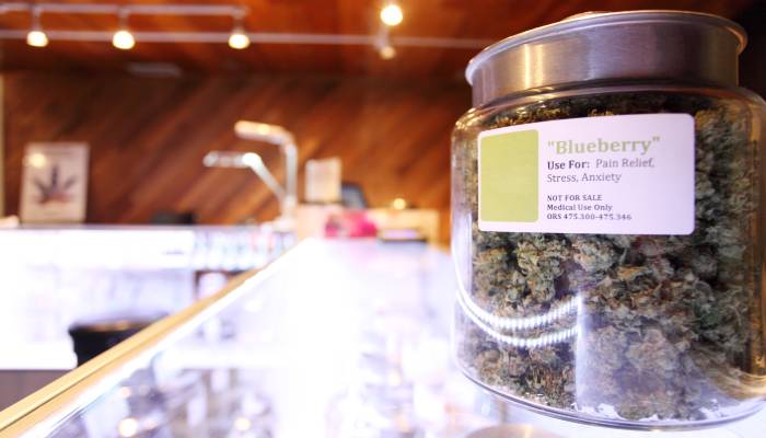 A close-up of a glass jar containing cannabis. The jar's label says "blueberry" and behind it is a wood paneled wall.