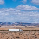 An overview of a white semi-truck driving through an empty highway in a dusty desert with mountains.