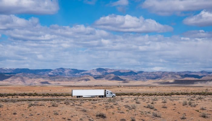 An overview of a white semi-truck driving through an empty highway in a dusty desert with mountains.