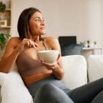 A relaxed, smiling woman eating out of a white bowl while lounging on a couch in athleisure wear.