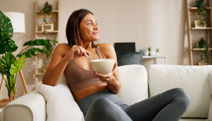 A relaxed, smiling woman eating out of a white bowl while lounging on a couch in athleisure wear.