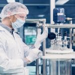 A scientist putting on rubber gloves. He is standing next to a hemp oil extraction machine that has a lot of metal parts.